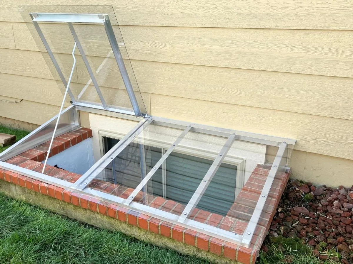 Window well cover propped open on brick well showing hinge and aluminum frame